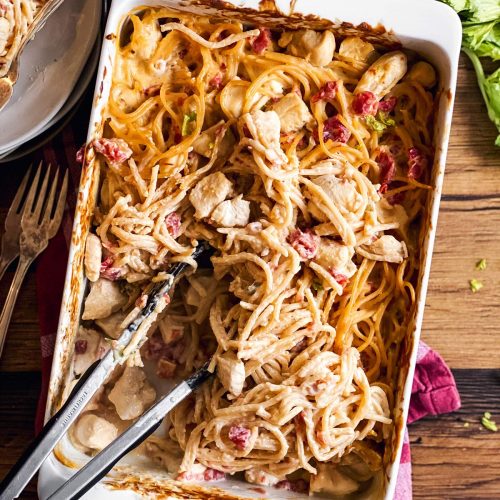 white casserole dish with chicken spaghetti and kitchen tongs on wooden background