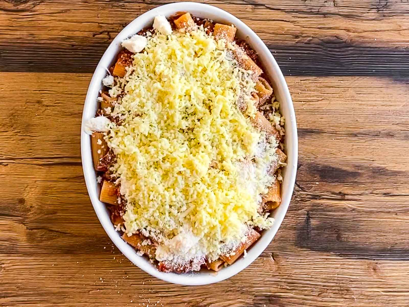 Rigatoni Bolognese covered with shredded cheeses in white casserole dish