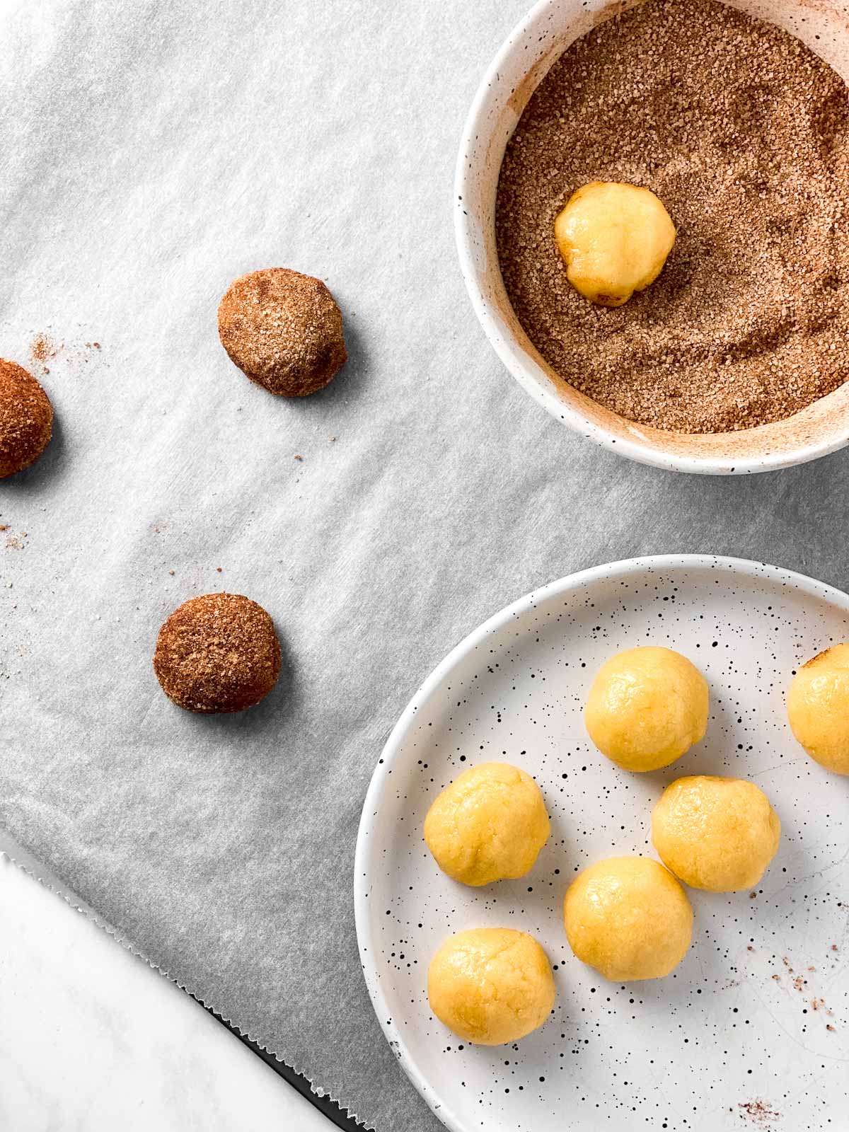 cookie dough balls on plate, one cookie dough ball in bowl with cinnamon sugar, three cookie dough balls on lined baking sheet