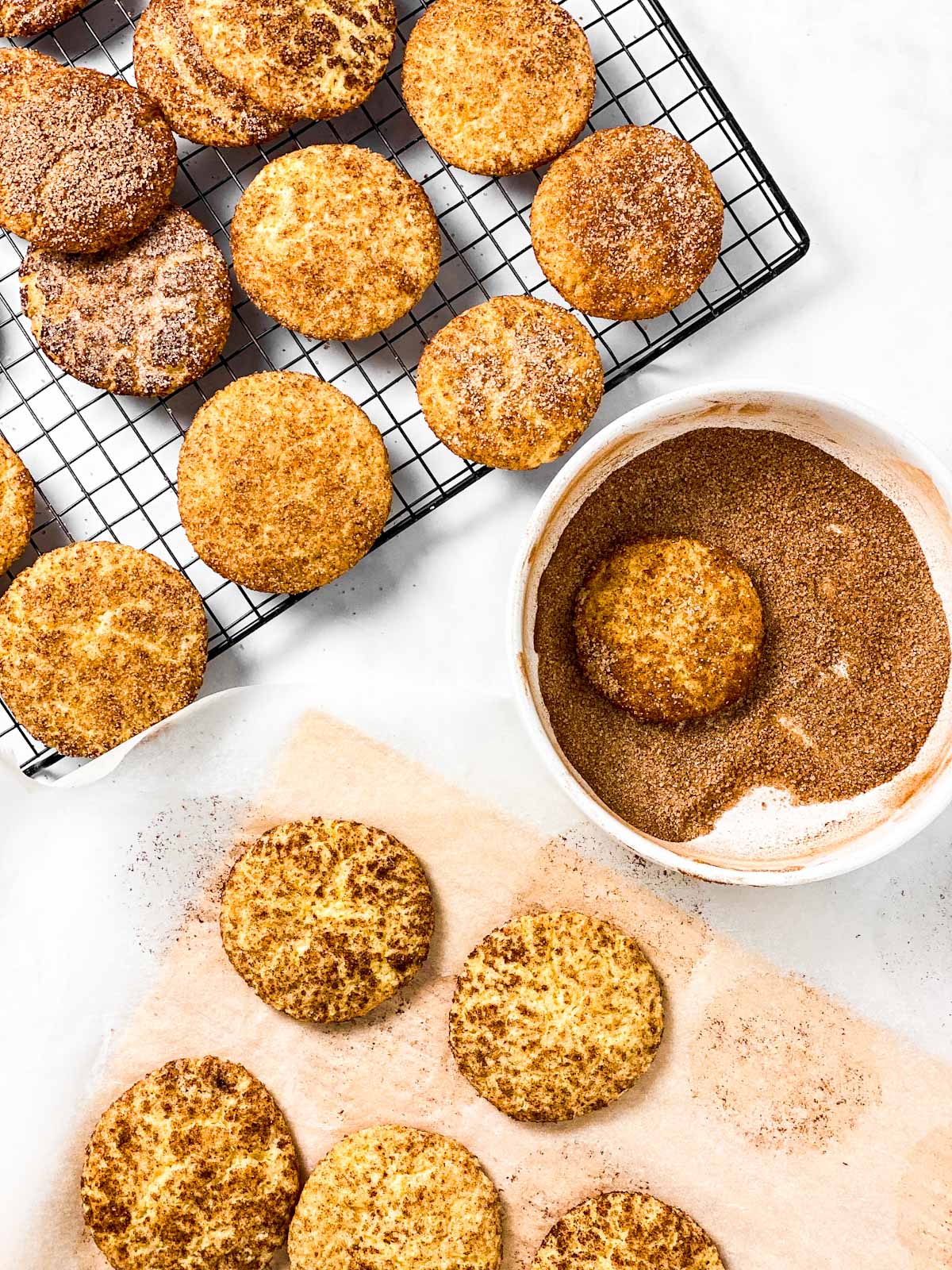 baked snickerdoodles on wire tray, one snickerdoodle in bowl with cinnamon sugar and five cookies on baking parchment