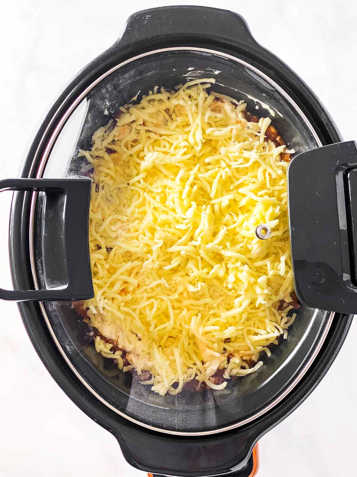slow cooker with closed lid, shredded mozzarella cheese visible underneath glass lid