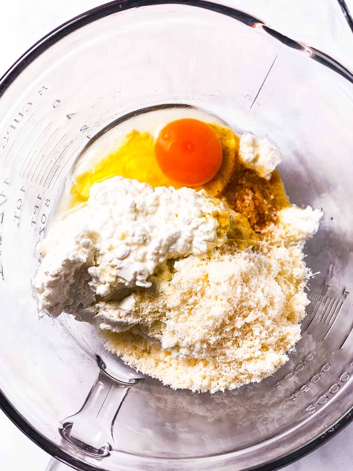 cracked egg and several different fresh and grated cheeses in glass bowl