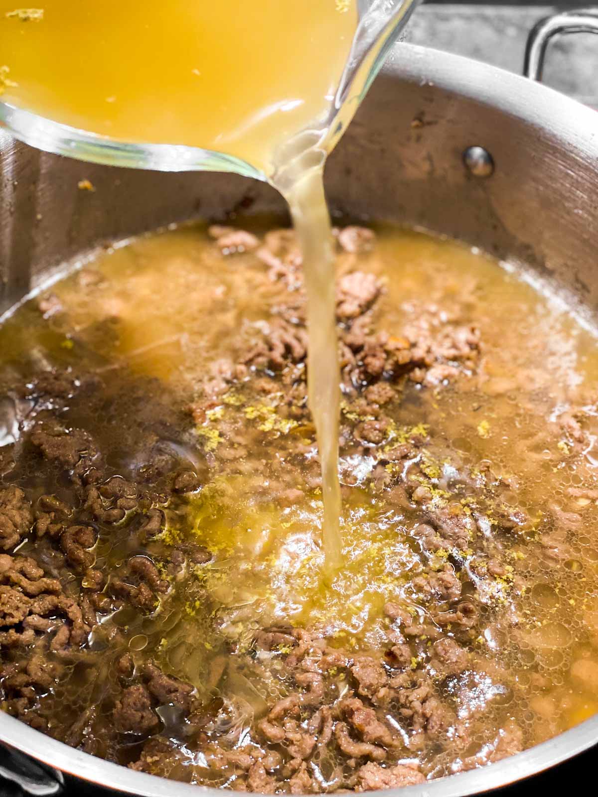 beef broth pouring from glass measuring jug into skillet with ground beef
