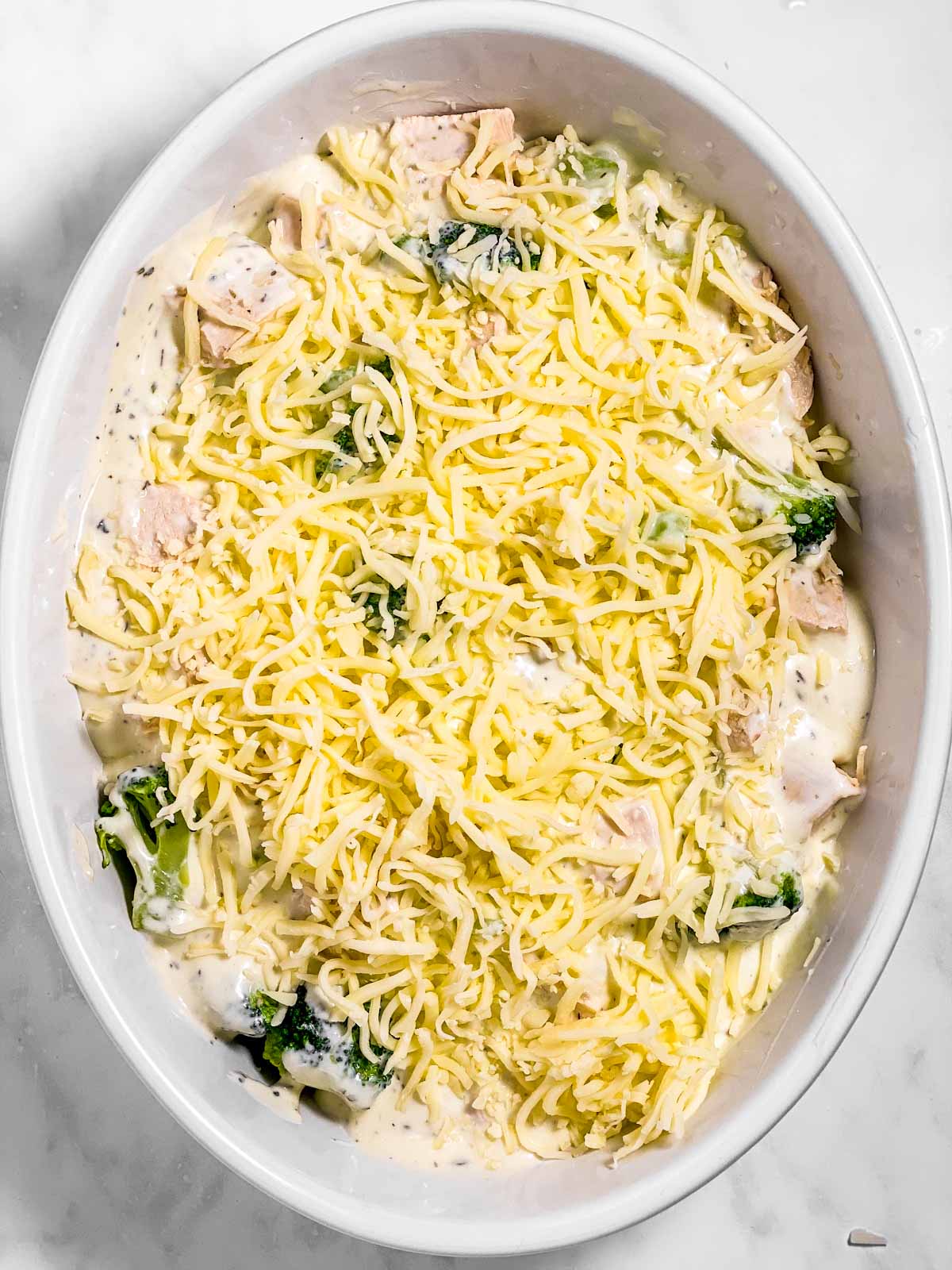 shredded mozzarella on top of chicken, broccoli and sauce in white oval casserole dish