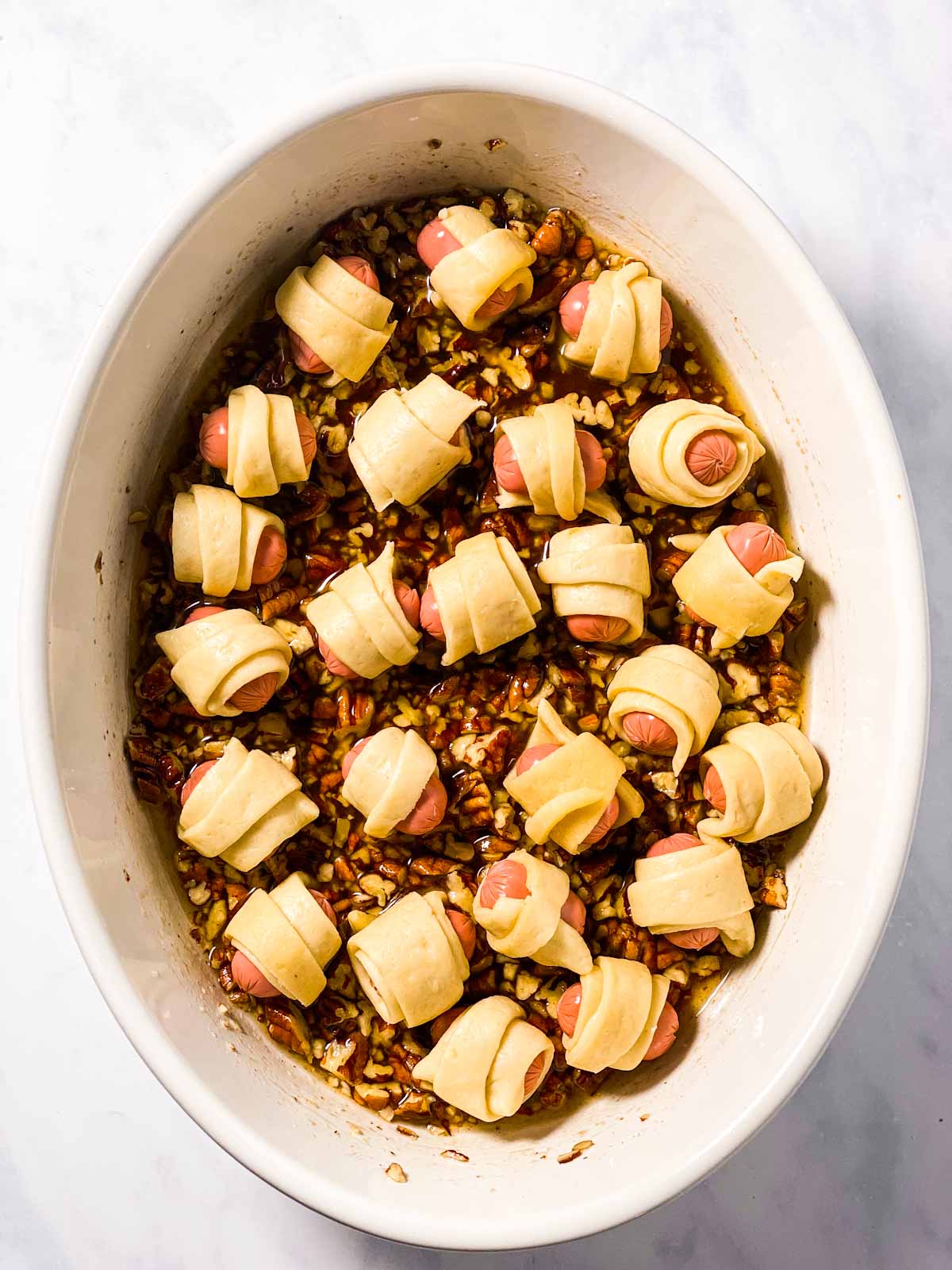 unbaked little smokies in crescent roll dough on top of pecan sugar mixture in white oval casserole dish