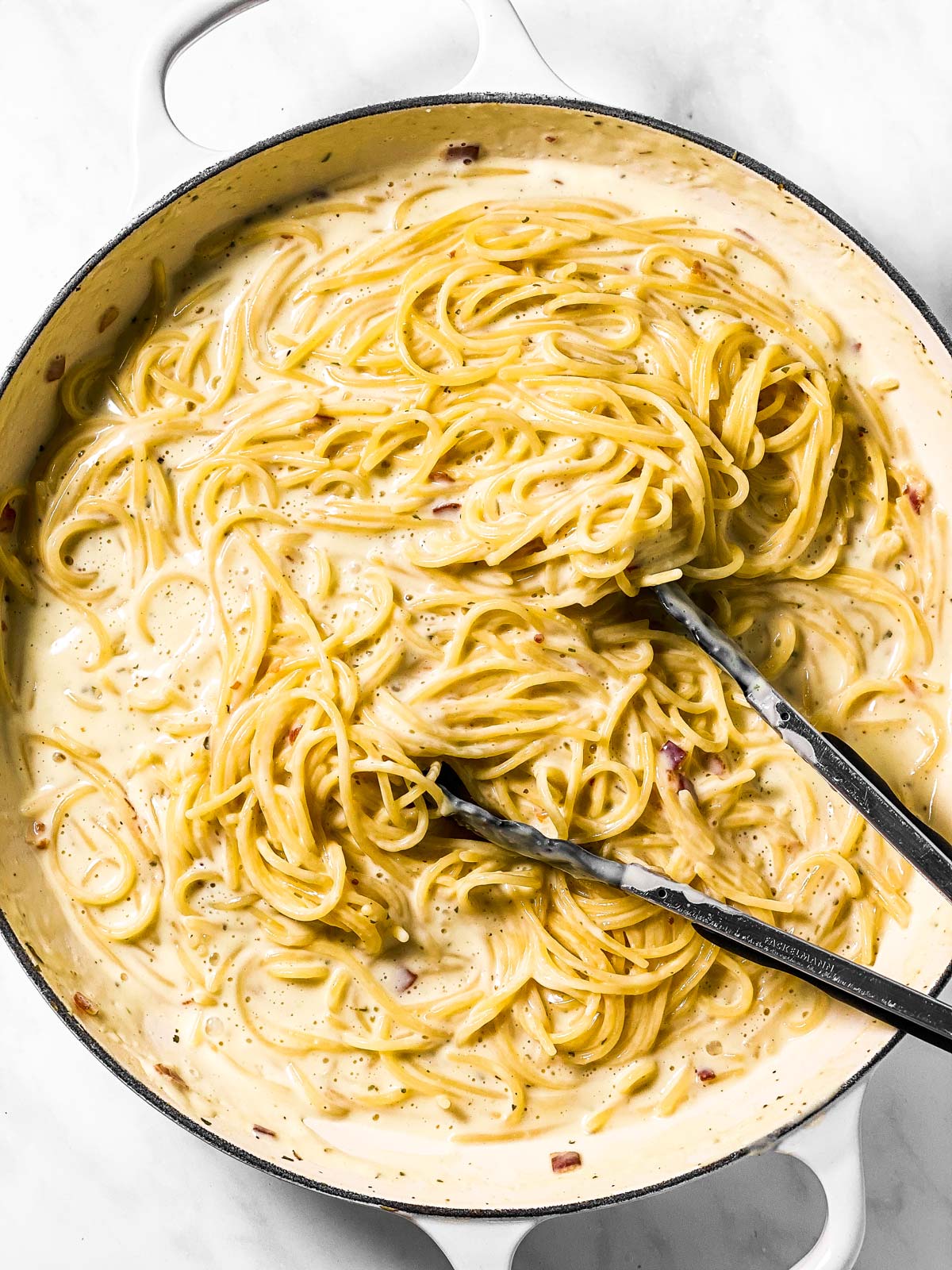 cooked spaghetti in creamy sauce in white skillet with kitchen tongs