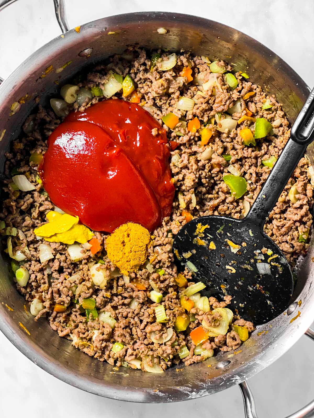 browned ground beef ad vegetables in skillet with ketchup, mustard, brown sugar and black cooking spoon